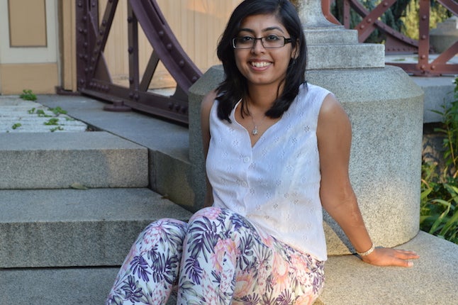 girl with dark hair and glasses sitting on grey stairs leaning against a cement column, wearing a pink shirt and multicolored palm tree patterned white pants 