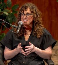 woman in glasses and black dress speaking into microphone