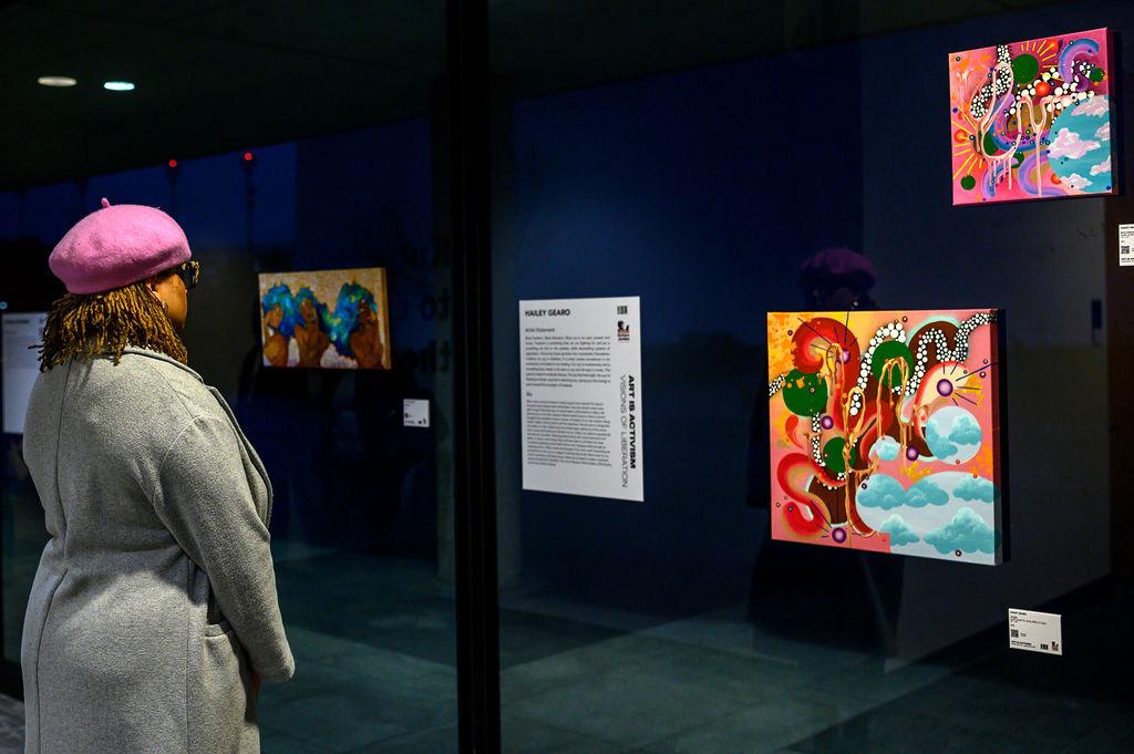 Art if Activism still #1 woman in pink hat looking at art works affixed to a glass windom