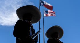 Two mariachi musicians stand in front of the flag posts on LBJ Plaza