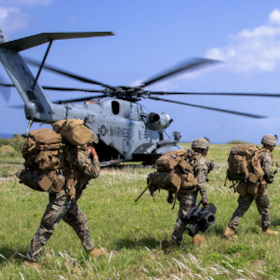 Soldiers wearing large pacs traverse a field, in front of a helicopter that likely just dropped them off