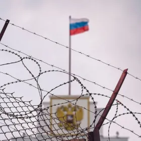 The image shows a barbed wire fence with the Russian flag in the background.