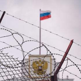 The image shows a barbed wire fence with the Russian flag in the background. 