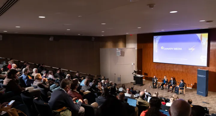 Crowd of people in lecture hall, watching 4 speakers on stage